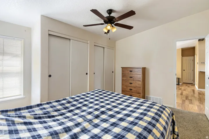 Spacious bedroom featuring triple sliding-door closets with mirrored panels, wood-finish dresser with multiple drawers, ceiling fan with integrated lighting, carpet flooring, and access to adjacent bathroom and hallway spaces