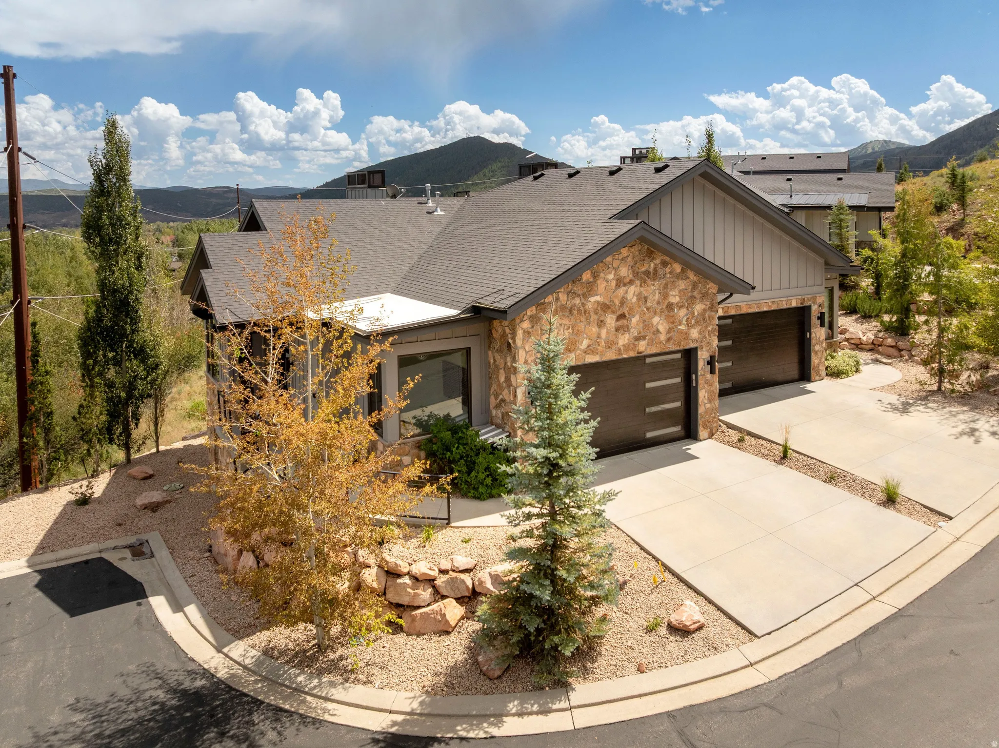 Multi-level roofline, stone and vertical siding exterior, two garage doors with horizontal accents, concrete driveway, and natural landscaping with mature trees