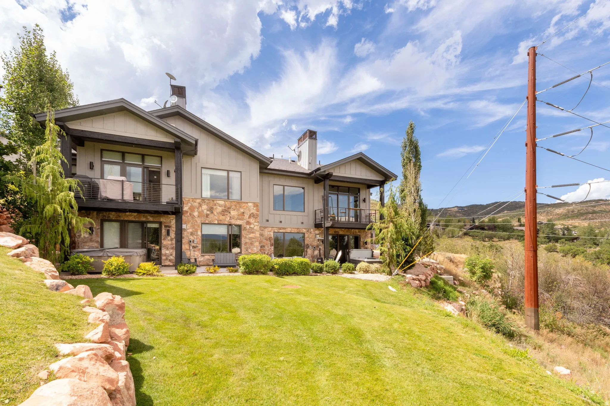 Stone and siding exterior with multiple balconies, a lower-level patio, a hot tub, and an expansive lawn