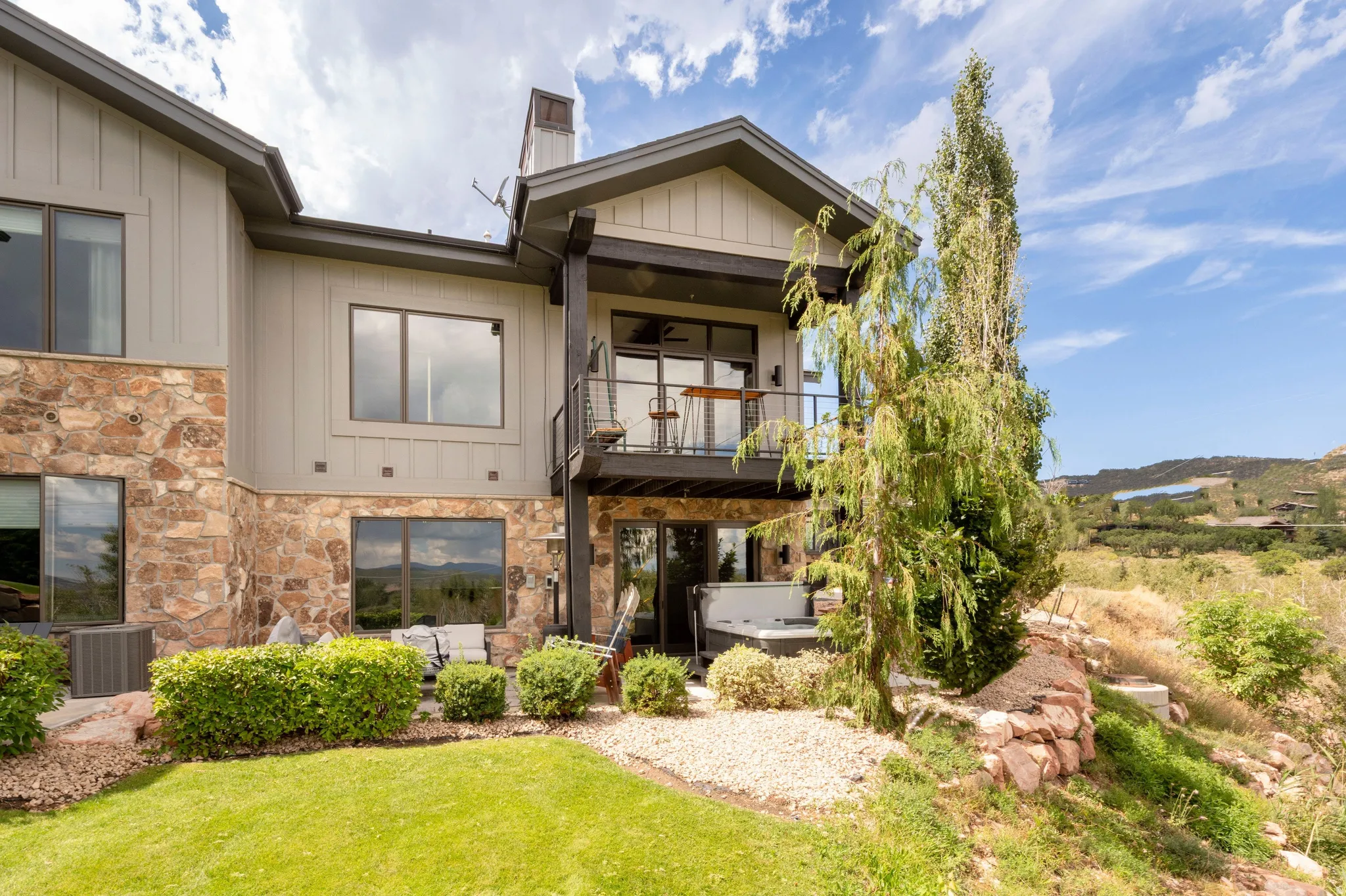 Stone and siding exterior with multiple windows, second-story balcony with metal railing, ground-level patio, and built-in hot tub