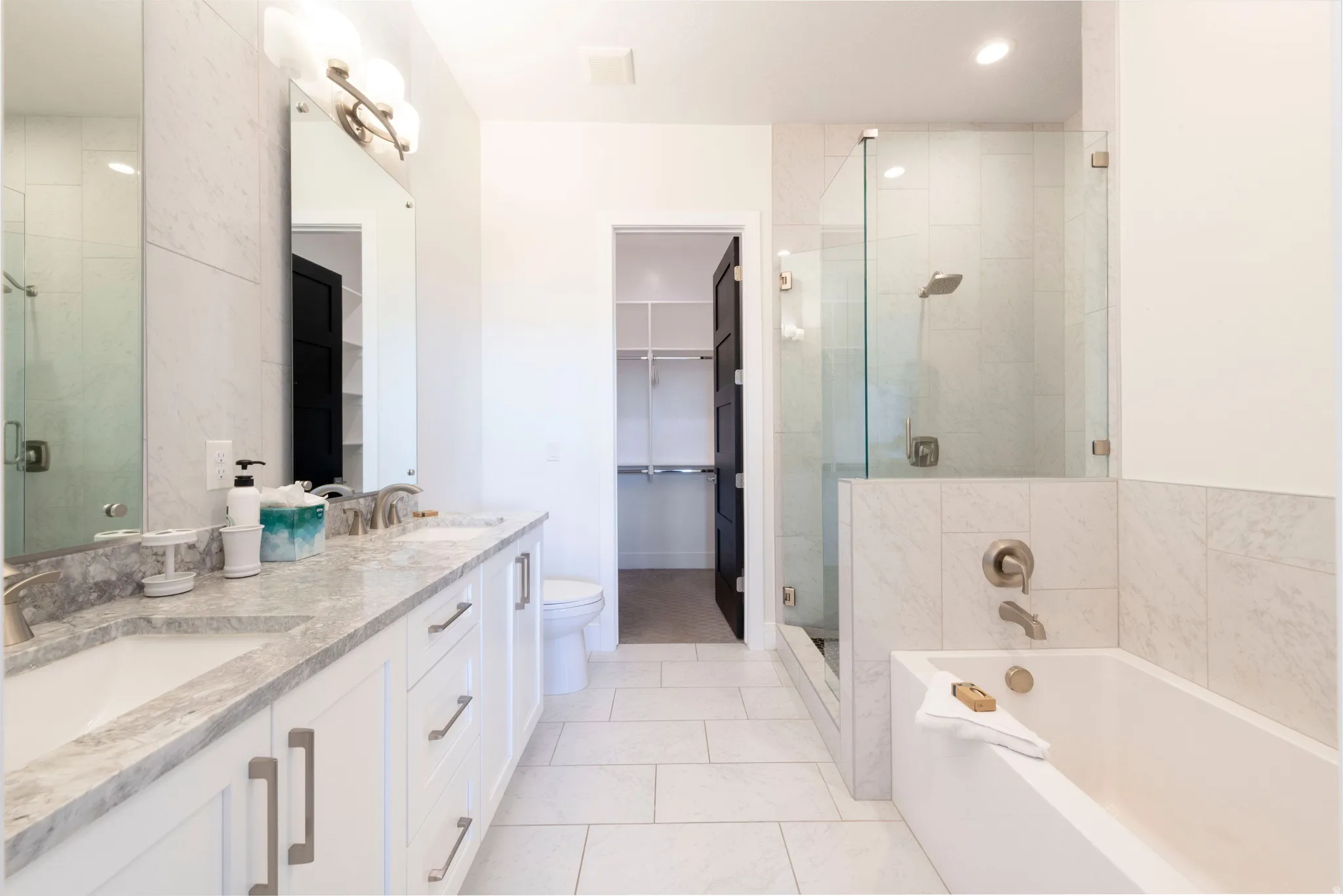 Dual vanity with undermount sinks and brushed nickel fixtures, glass-enclosed shower with tiled surround, built-in soaking tub, and tile flooring