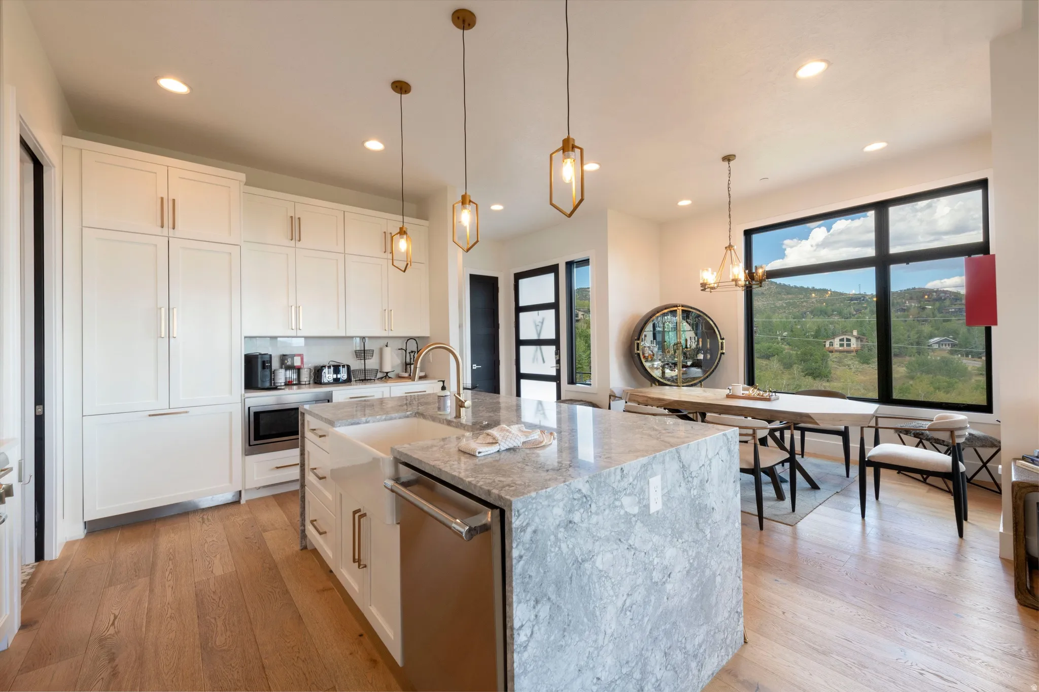 White cabinetry with gold-tone hardware, wood-finish flooring, a waterfall kitchen island with an undermount sink, and large picture windows