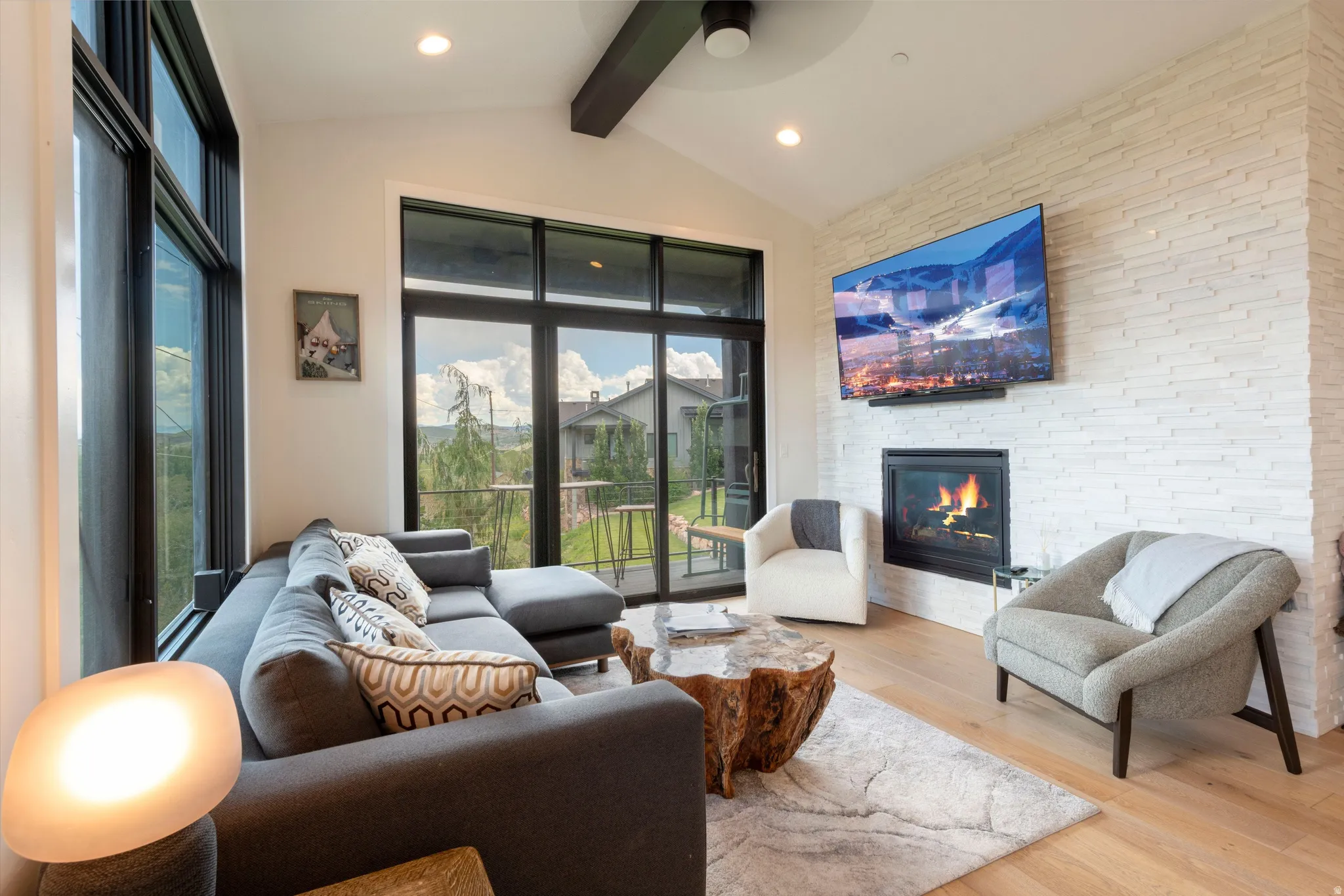 Vaulted ceiling with exposed beam, floor-to-ceiling windows, built-in gas fireplace, light wood-finish flooring, and a stacked stone accent wall