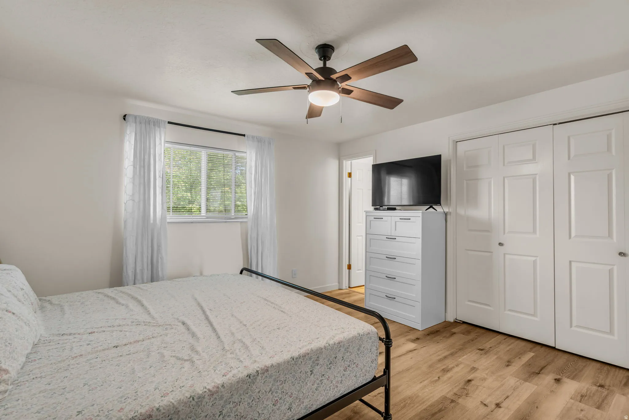 Bright secondary bedroom featuring hardwood flooring, a ceiling fan with integrated lighting, and ample natural light from a multi-pane window with sheer curtains