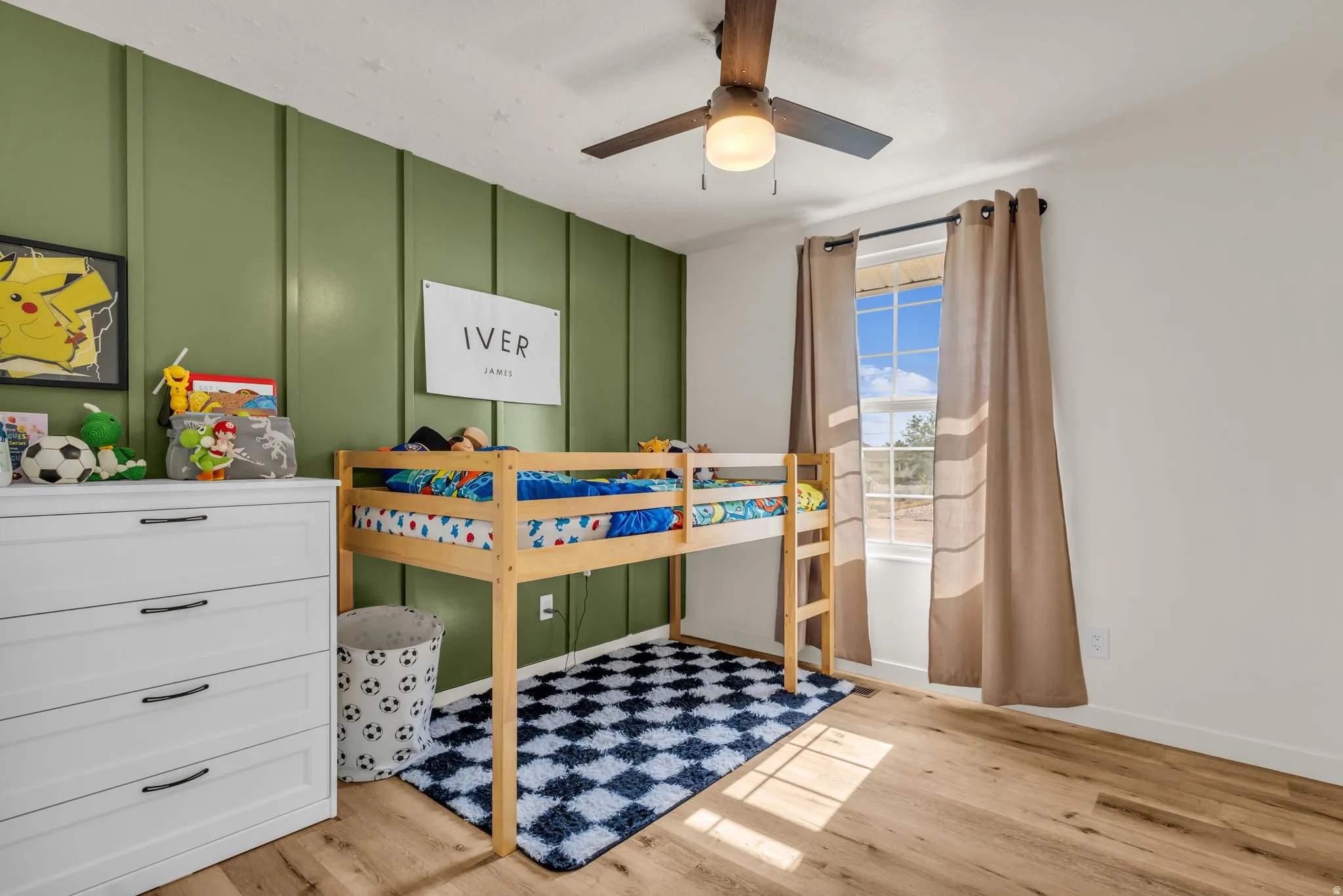 Wood-finish flooring, accent wall with vertical paneling, single window with trim, ceiling fan with integrated lighting, and white walls