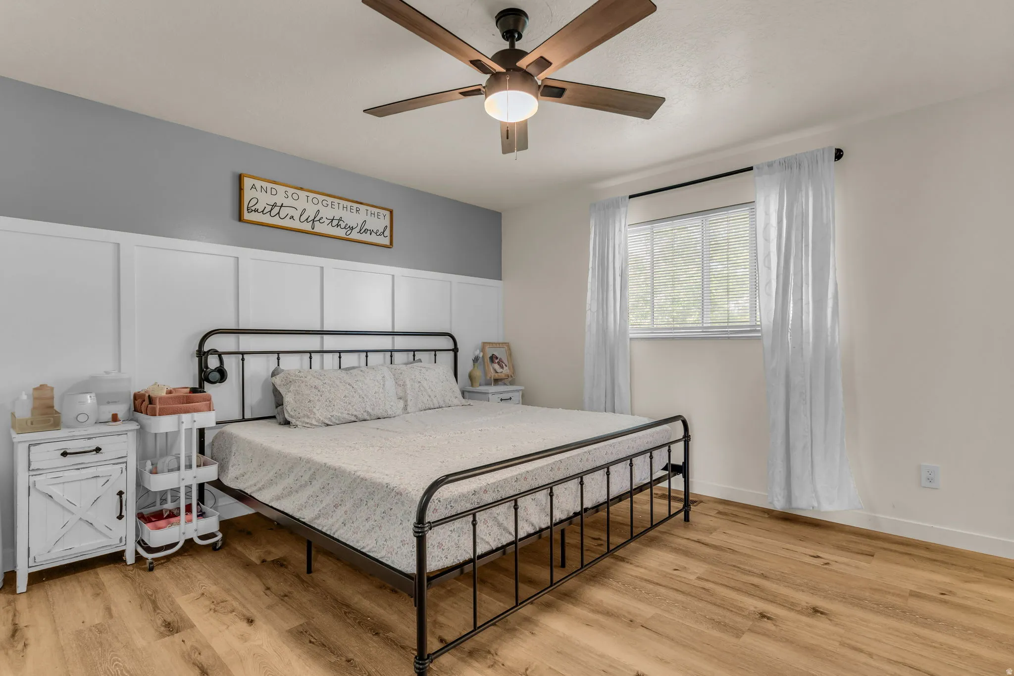 Wood-finish flooring, ceiling fan with integrated lighting, window with blinds, and white wainscoting accent wall