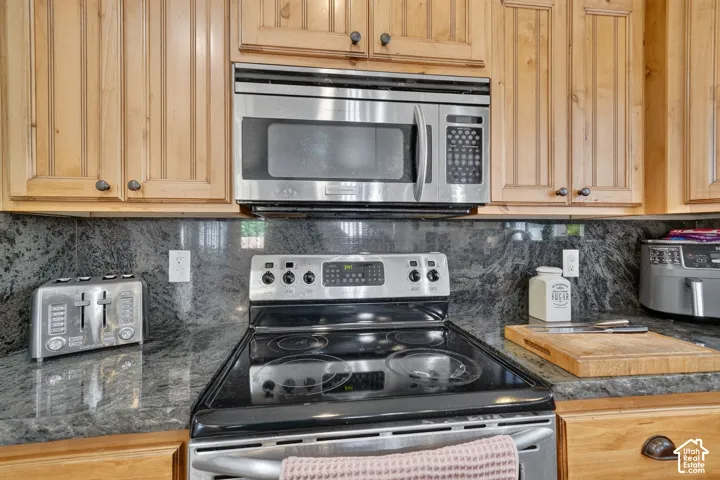 Stainless steel microwave, stainless steel range, wood cabinetry, stone countertops, and a stone backsplash