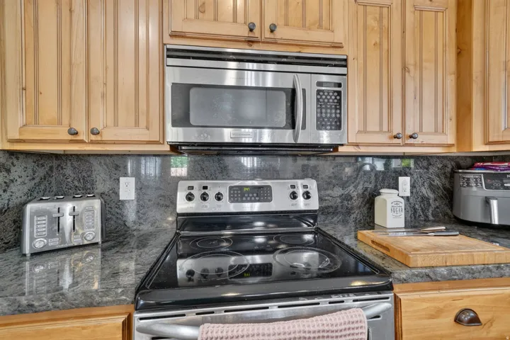 Stainless steel microwave, stainless steel range, wood cabinetry, stone countertops, and a stone backsplash