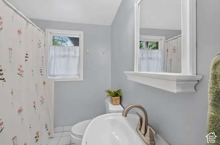 White tiled flooring, white-framed window, white-framed mirror with shelf, white toilet, and a brushed nickel-finish faucet