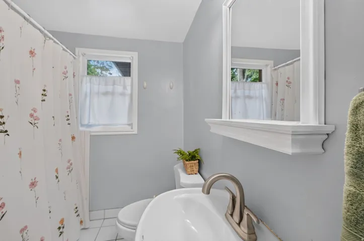 White tiled flooring, white-framed window, white-framed mirror with shelf, white toilet, and a brushed nickel-finish faucet