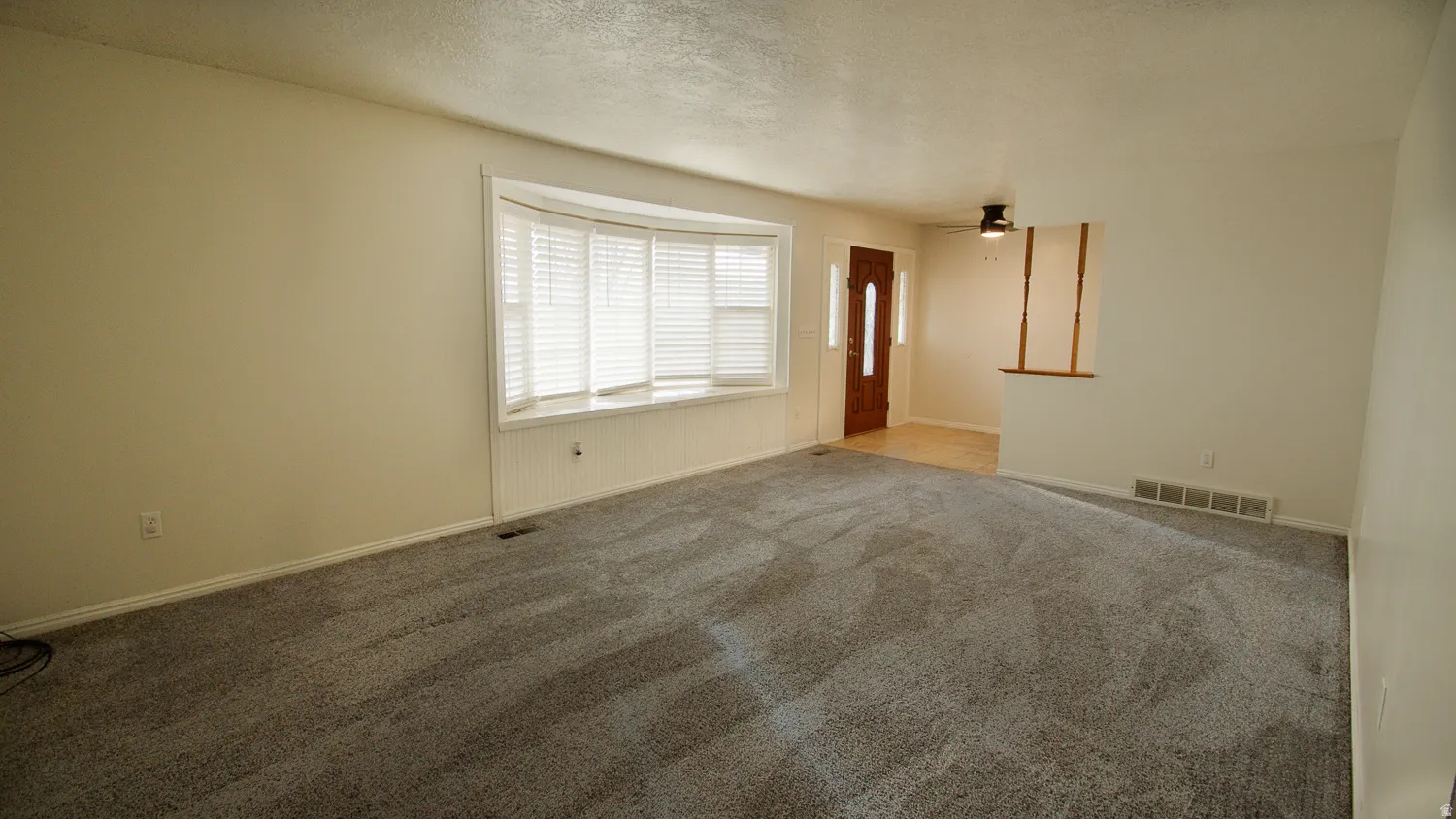 Spacious room featuring gray carpeting, a bay window with white blinds, a wood-paneled entry door, and a ceiling fan