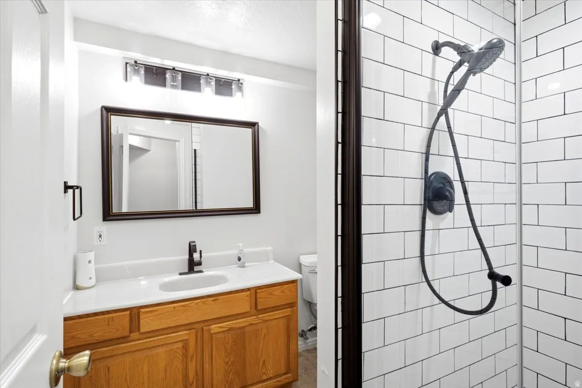 Modern primary bathroom featuring a wood-finish vanity with dual sinks, framed mirror with integrated lighting, and subway tile shower enclosure with rainfall showerhead and handheld spray fixture