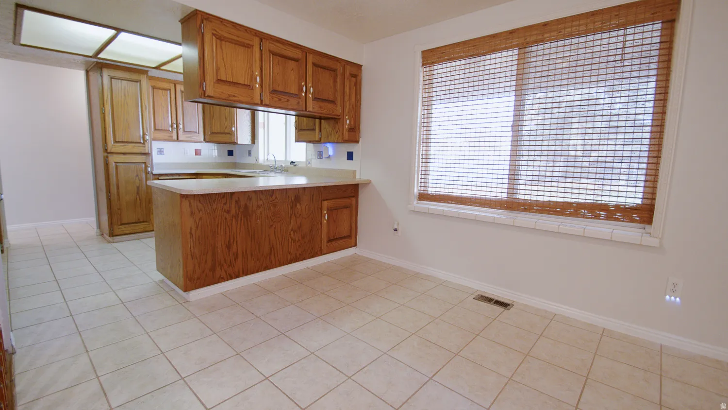 Spacious open-concept kitchen and dining area featuring wood-finish cabinetry, tile flooring throughout, and abundant natural light from large windows with horizontal blinds