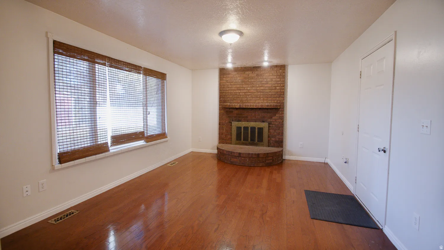 Spacious living room showcasing rich wood-finish flooring, a brick fireplace with dark wood mantel, large multi-pane windows with interior blinds providing natural light, and white painted doors and trim throughout