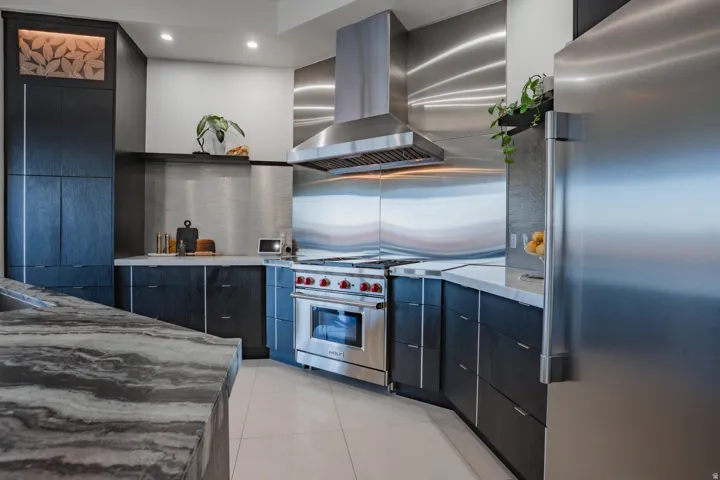 Kitchen featuring stainless steel appliances, dark wood-finish cabinetry, patterned stone countertops, and a full-height stainless steel backsplash