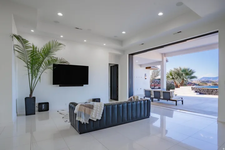 White large format tile flooring, recessed ceiling lighting, a wall-mounted entertainment fixture, and expansive sliding glass doors opening to an outdoor patio with a pool
