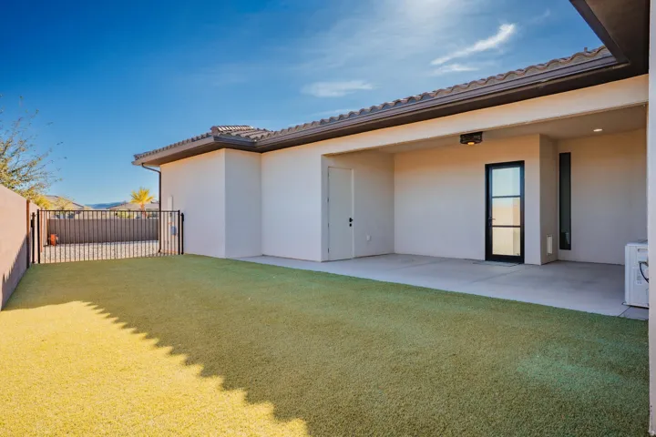 Expansive artificial turf yard with a covered patio, stucco exterior, black metal fencing, and a modern glass-paneled door