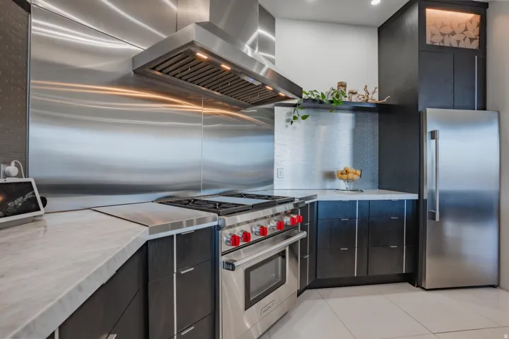 Stainless steel range hood with integrated lighting, full stainless steel backsplash, professional-grade range, dark wood-finish cabinetry, and light-toned countertops