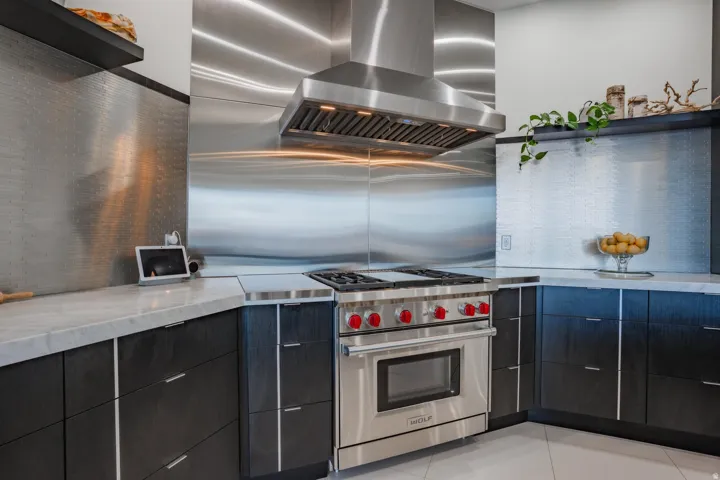 Stainless steel range hood with integrated lighting, professional-grade gas range, full stainless steel backsplash, dark wood-finish cabinetry, and light-toned countertop