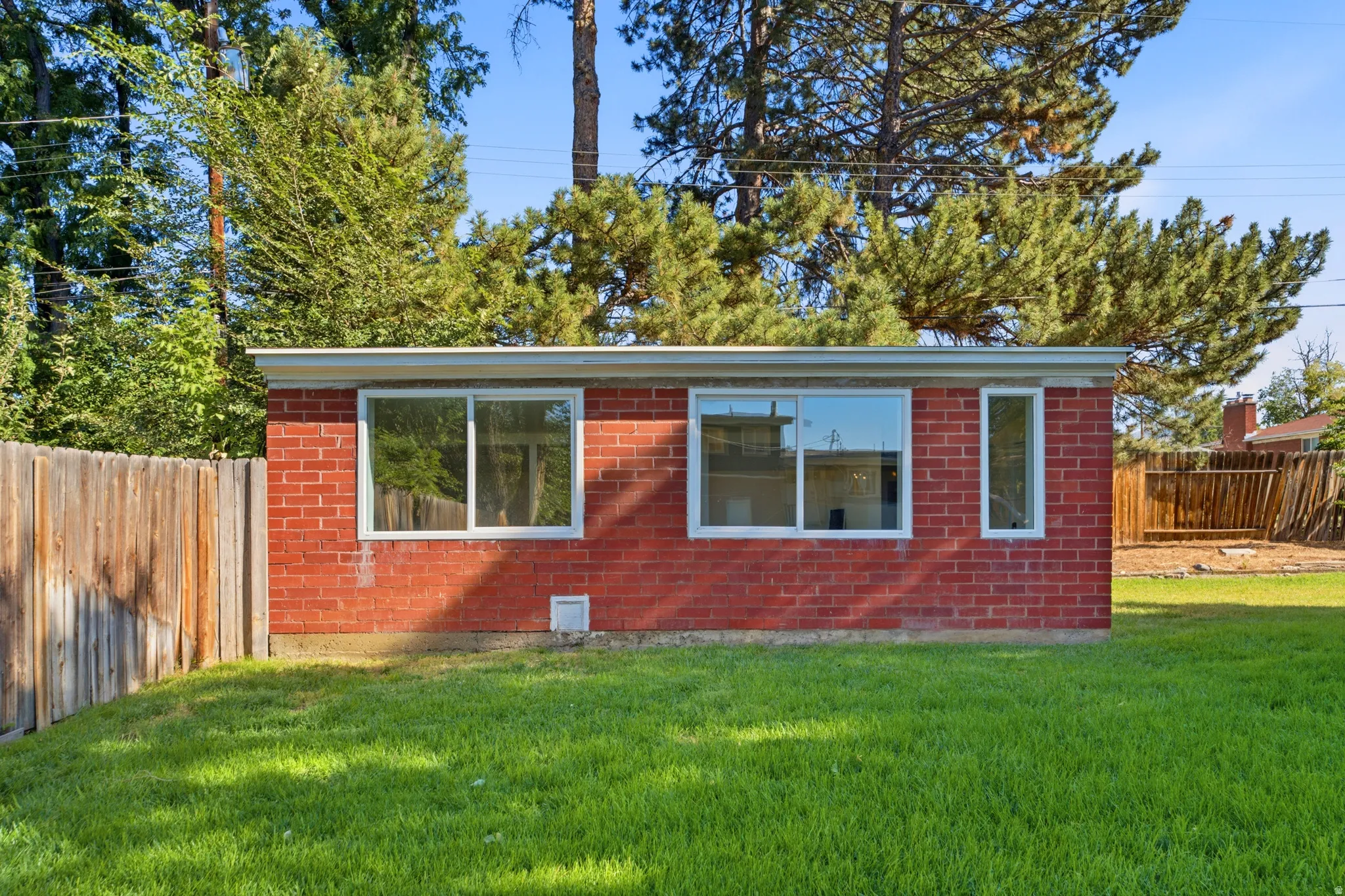 Brick exterior home featuring expansive windows with white trim, manicured lawn, mature evergreen trees, wooden privacy fencing, and open grounds ideal for outdoor use