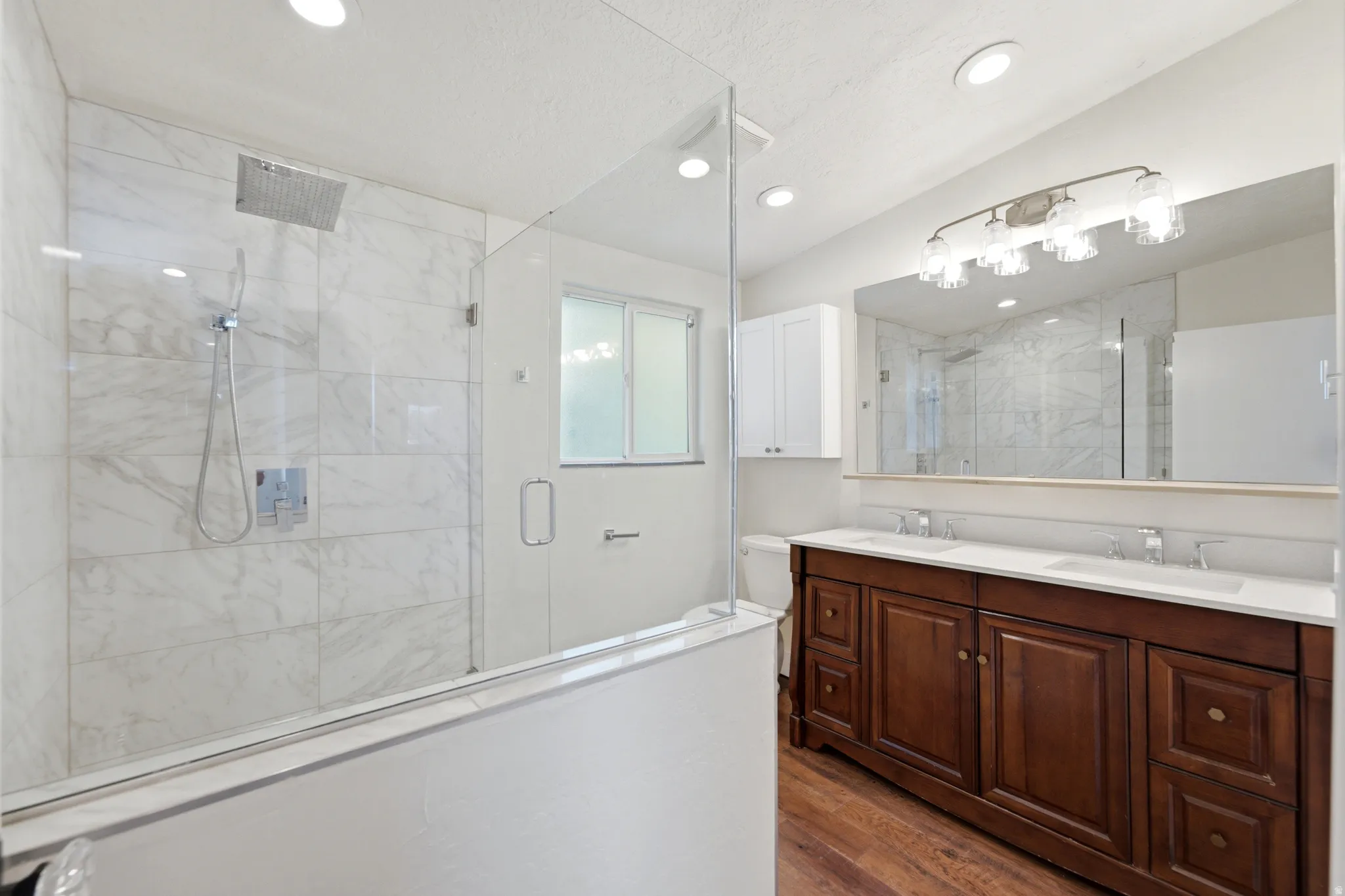 Spacious bathroom featuring a double vanity with wood-finish cabinetry, a large vanity mirror, and a glass-enclosed shower with marble-look tile