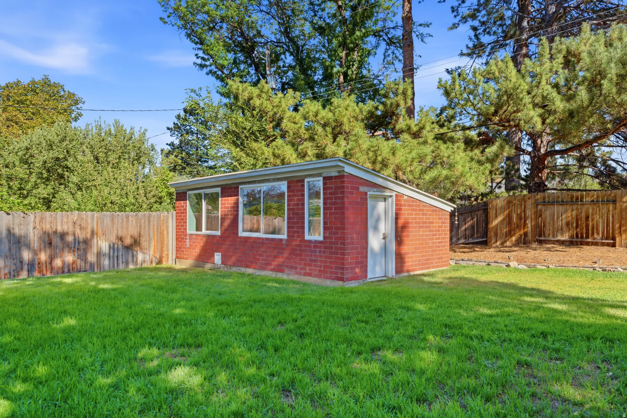 Brick auxiliary structure with multiple windows, a solid entry door, and a flat roof, set within a fenced yard featuring mature trees and a lawn area
