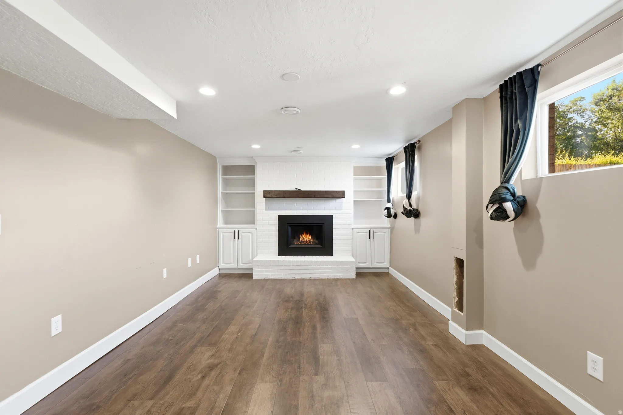 Recessed lighting illuminating wood-finish flooring, a white brick fireplace with a wood mantel, and built-in shelving with lower cabinetry