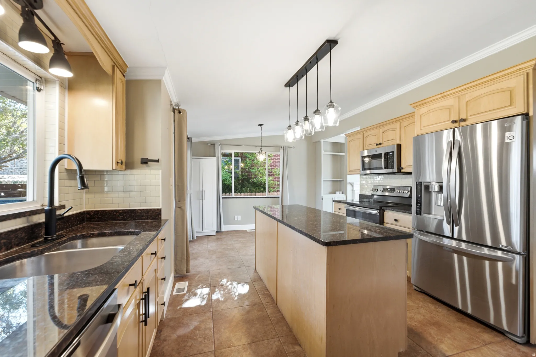 Kitchen featuring light wood cabinetry, stainless steel appliances, dark stone countertops, a tile backsplash, and a central island with pendant lighting