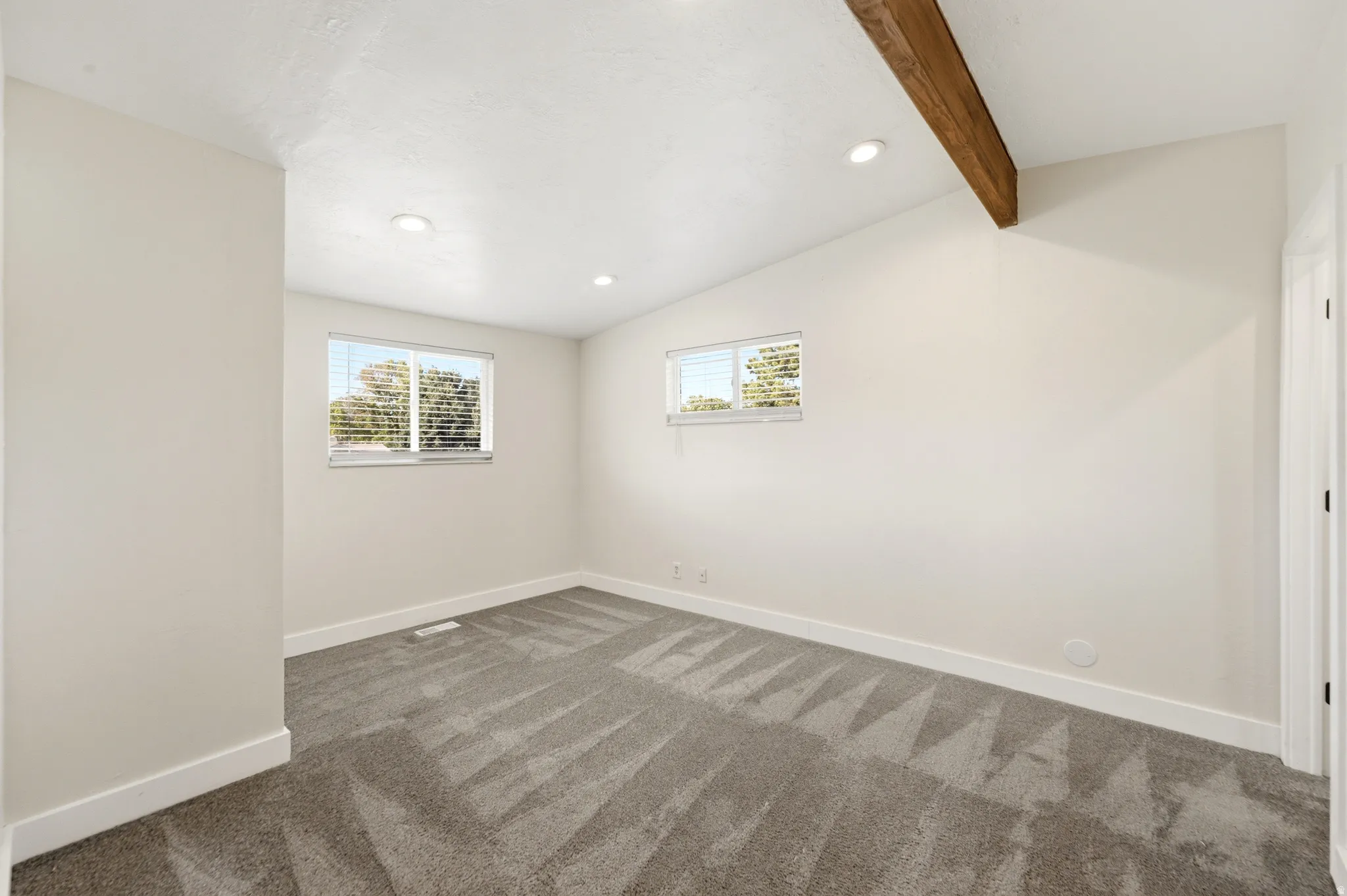 Recessed lighting, wood beam ceiling accent, light gray patterned carpet, two windows with blinds, and white trim