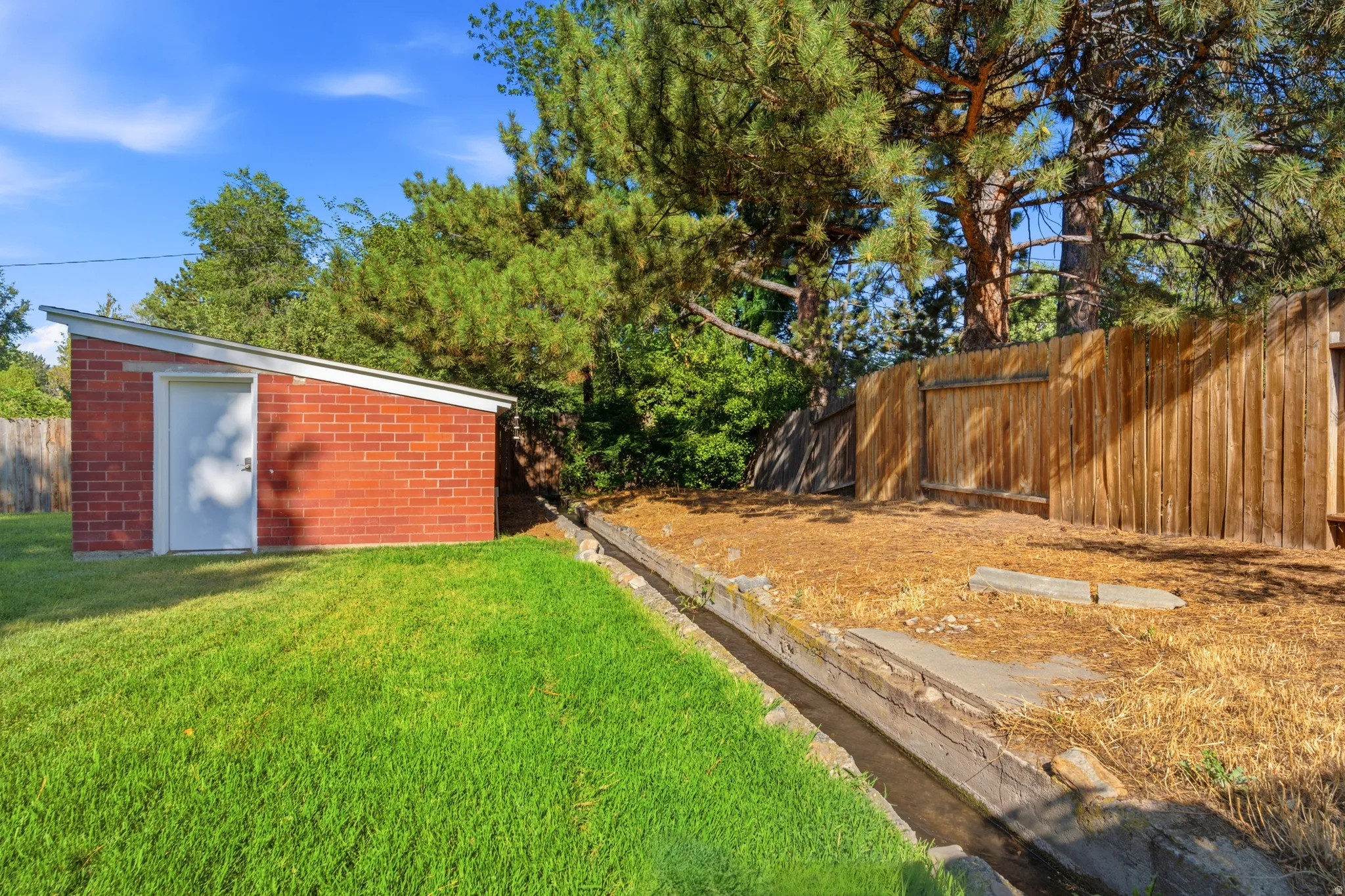 Red brick storage shed with a white door, sloped roof, and lush green lawn