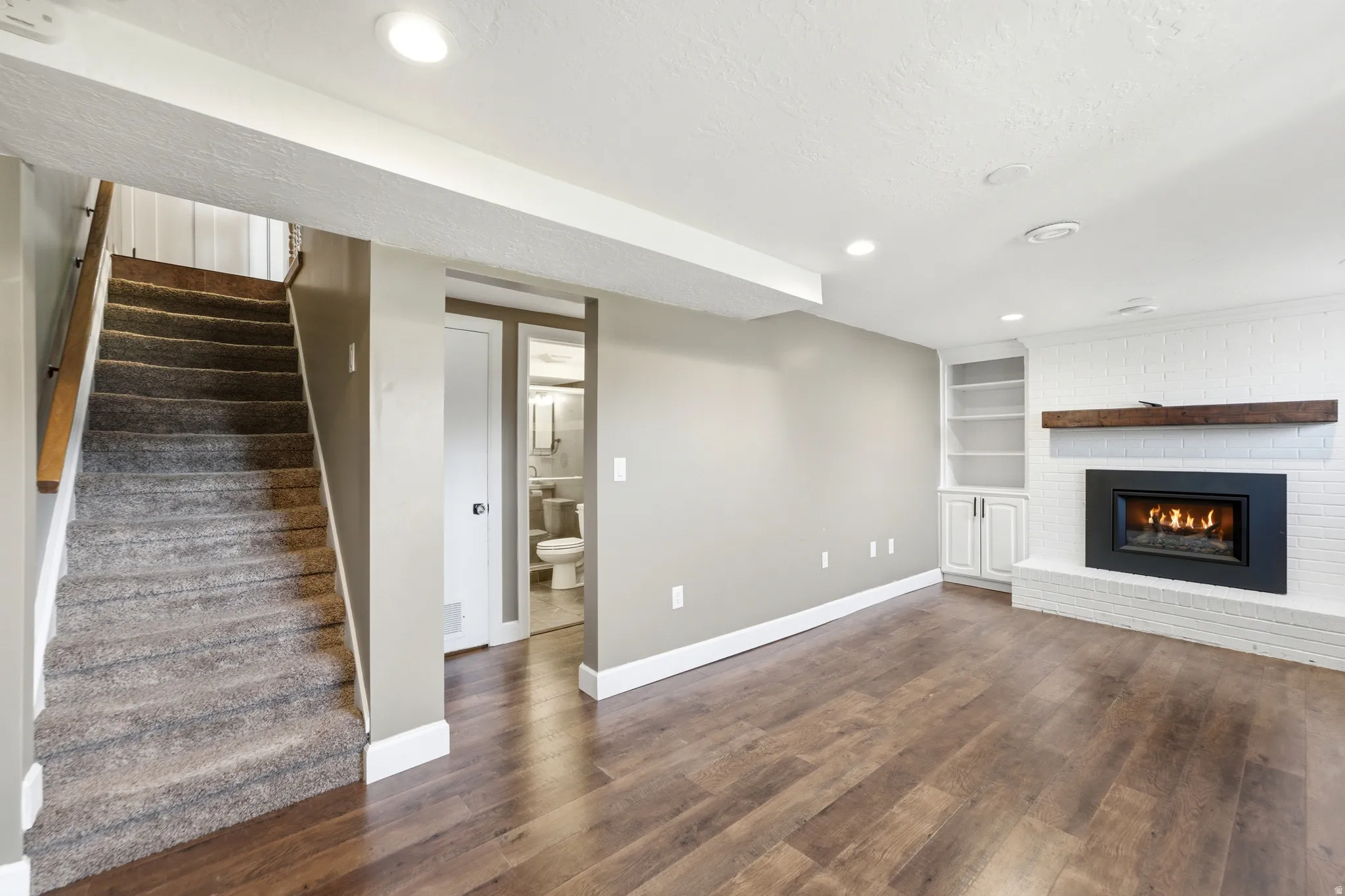 Spacious living area featuring wood-finish flooring, a white brick fireplace with a wood mantle, built-in shelving, and recessed lighting
