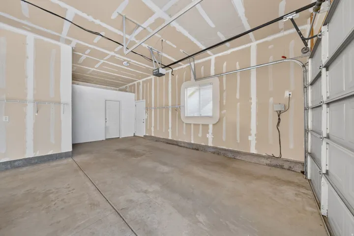 Spacious garage with concrete flooring, overhead garage door with automatic opener hardware, interior service door, and exposed ceiling framing with integrated lighting infrastructure