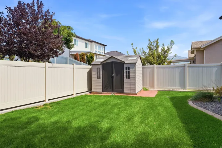 Expansive grass lawn, fully fenced with light-toned privacy fencing, featuring a storage shed with dual doors and an adjacent paver patio