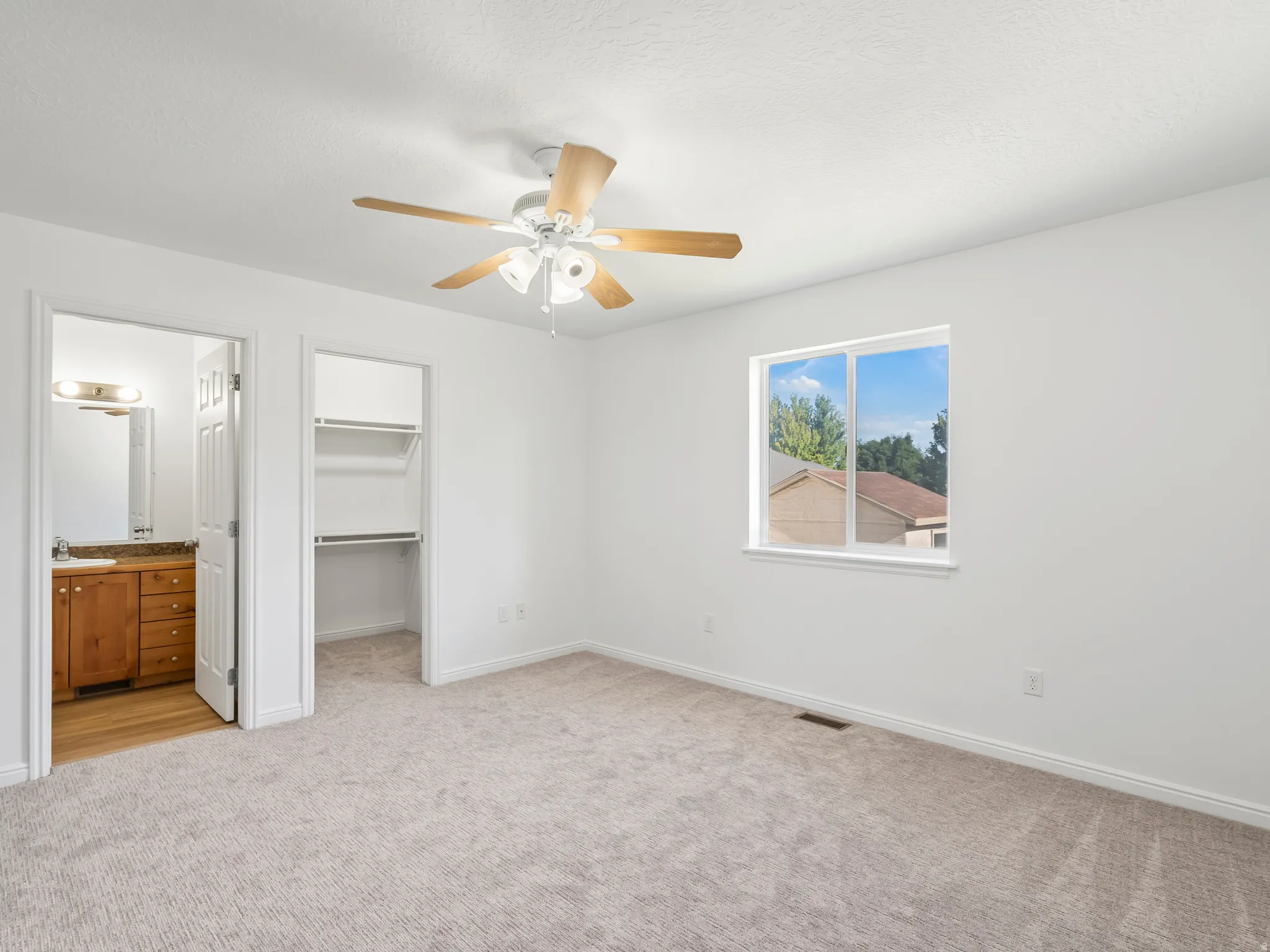 Recessed window with exterior scenery, overhead ceiling fan with light fixture, wall-to-wall carpeting, built-in closet shelving, and an adjacent bathroom with a wood-finish vanity and mirror