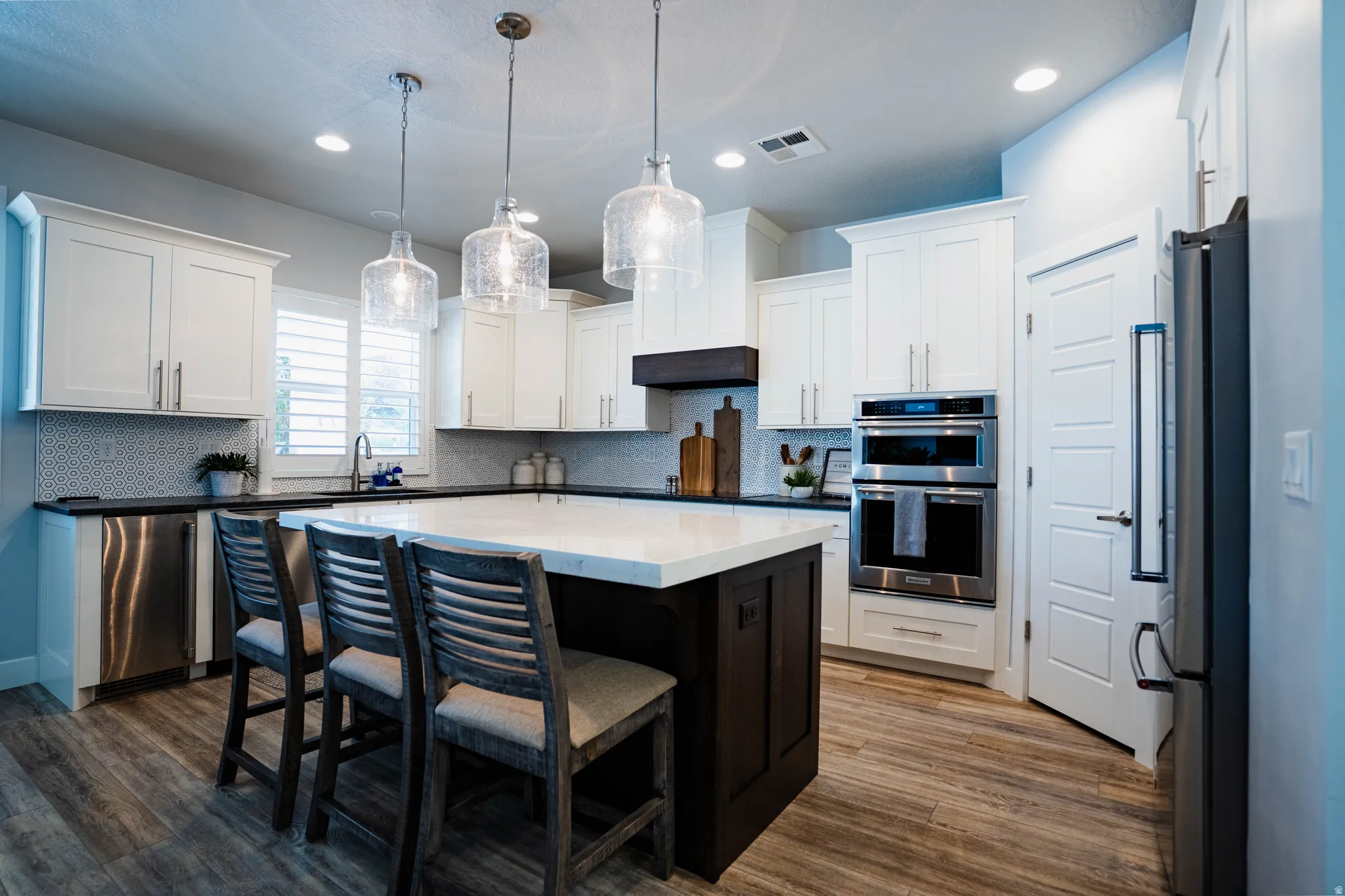 Bright kitchen featuring white shaker cabinetry, dark countertops, wood-finish flooring, stainless steel appliances, and a central island with counter seating