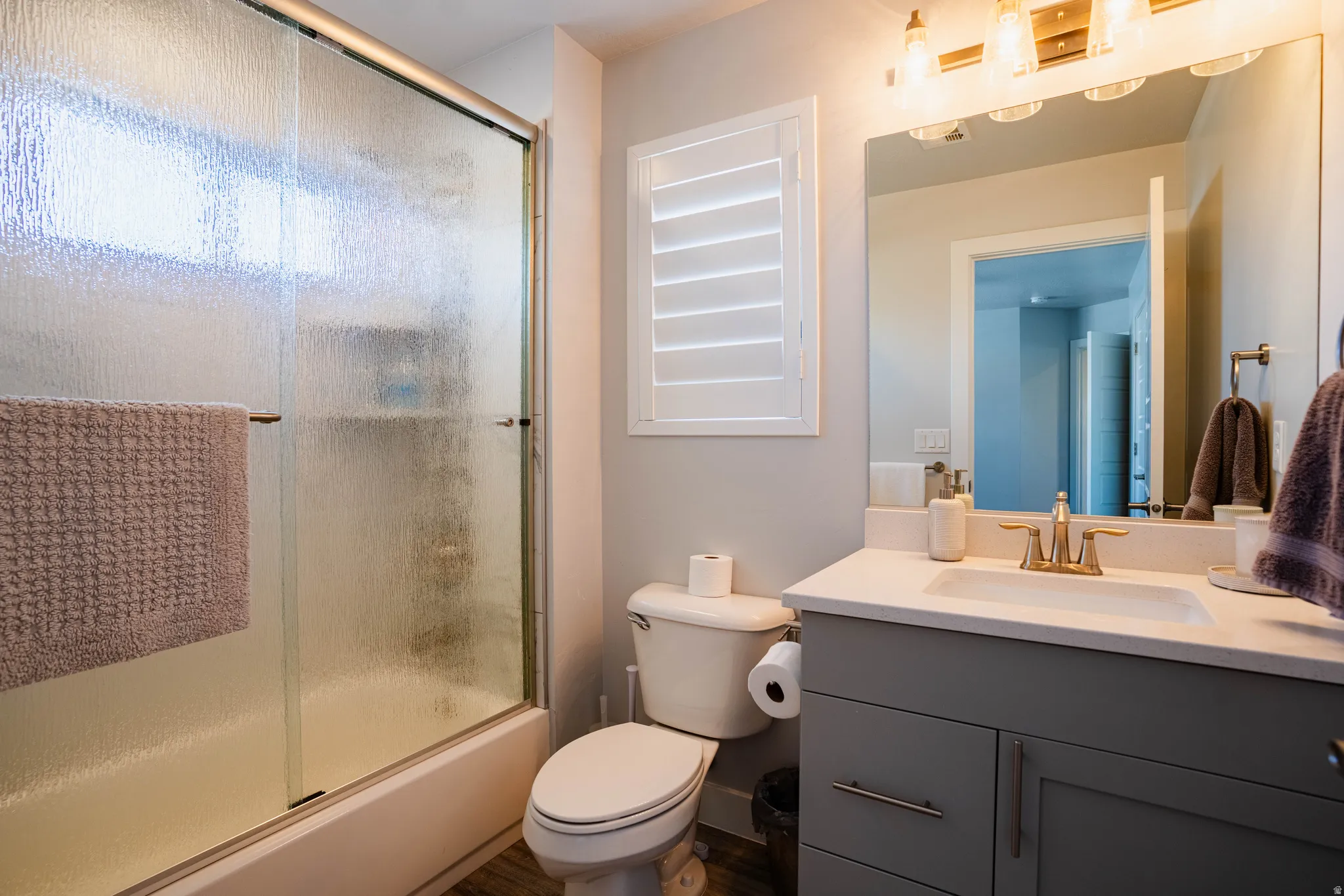 Wood-finish flooring, sliding glass shower enclosure, white vanity with integrated sink, gold-toned faucet, and full-width mirror