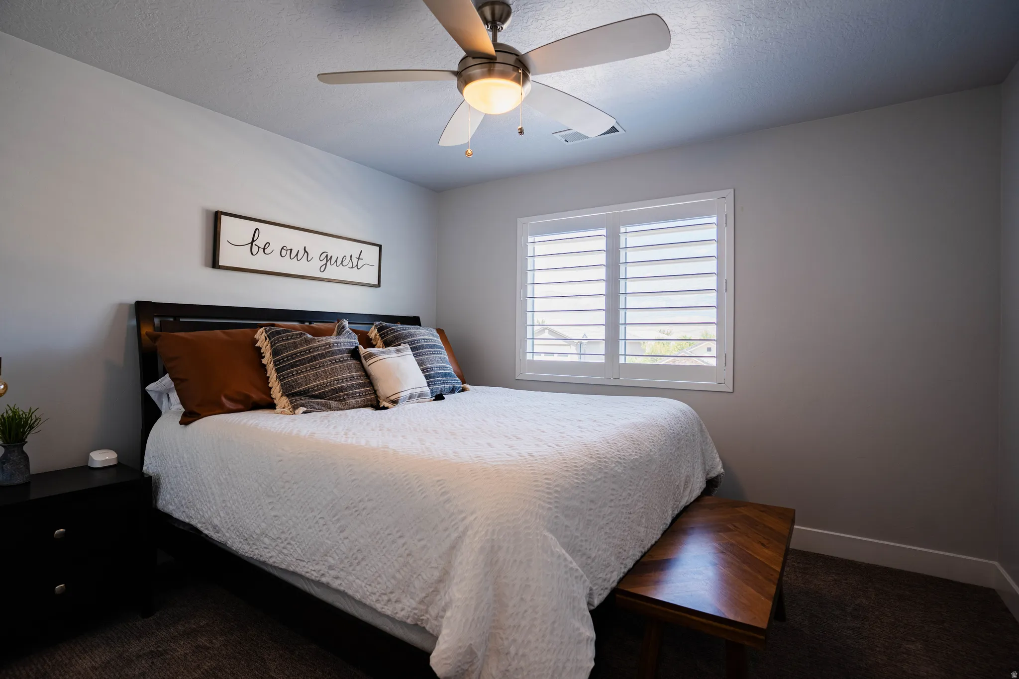 Ceiling fan with integrated lighting, window with white plantation shutters, dark wood-finish bed frame, dark-toned carpet flooring, and light gray wall paint