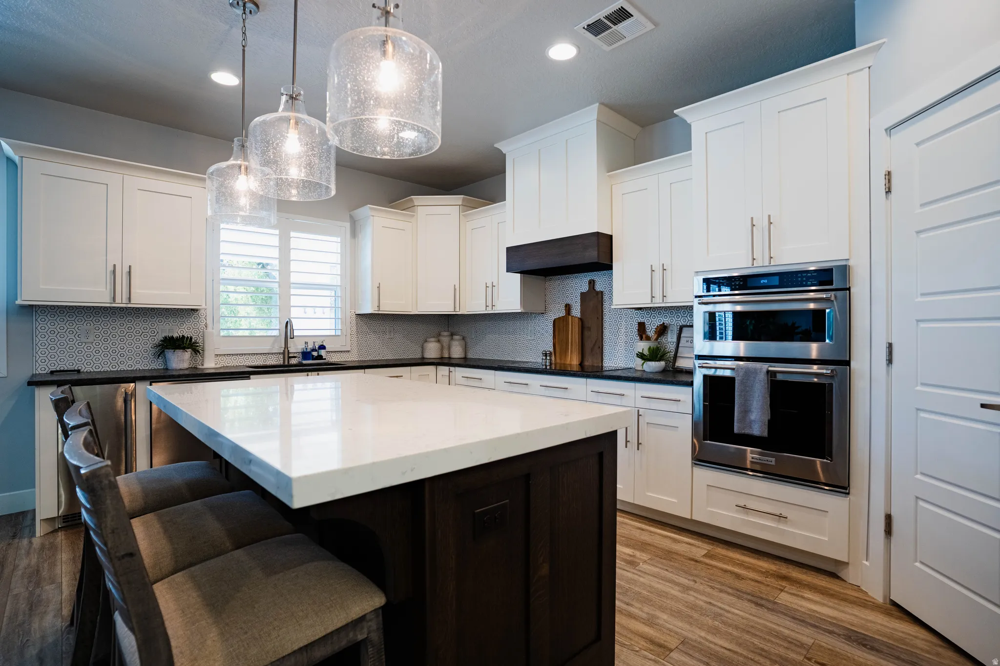 Kitchen featuring white shaker cabinetry, stainless steel double ovens, a large center island with seating, a dark wood-finish hood, and wood-finish flooring