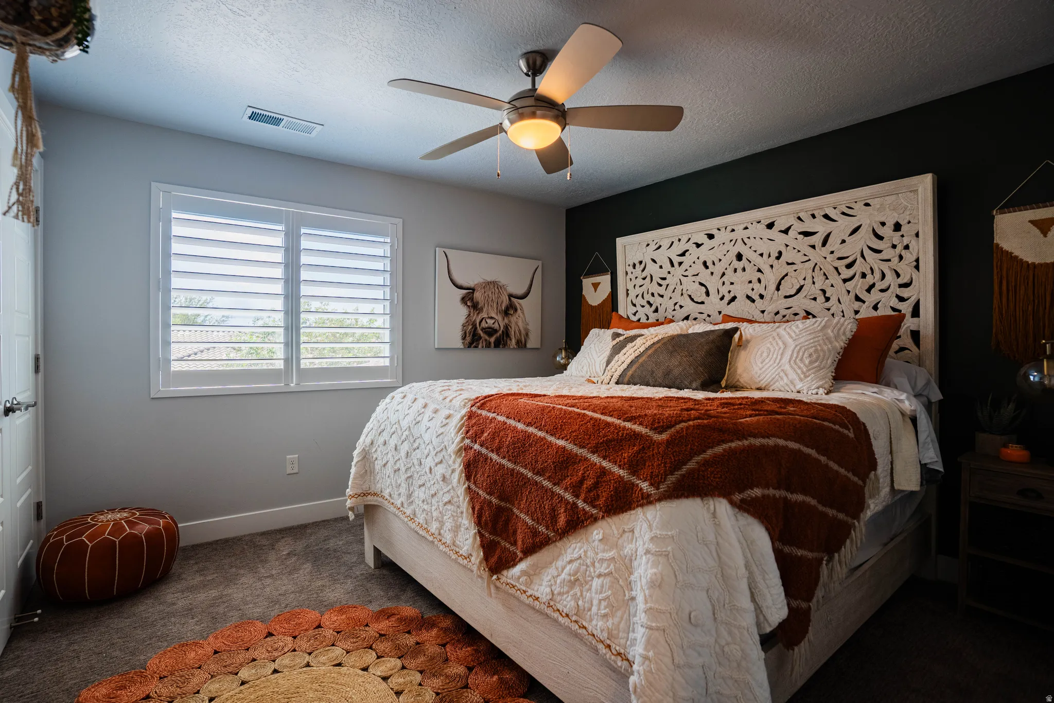 Window with white louvered shutters, ceiling fan with integrated lighting, dark accent wall, textured carpet flooring, and a carved decorative headboard