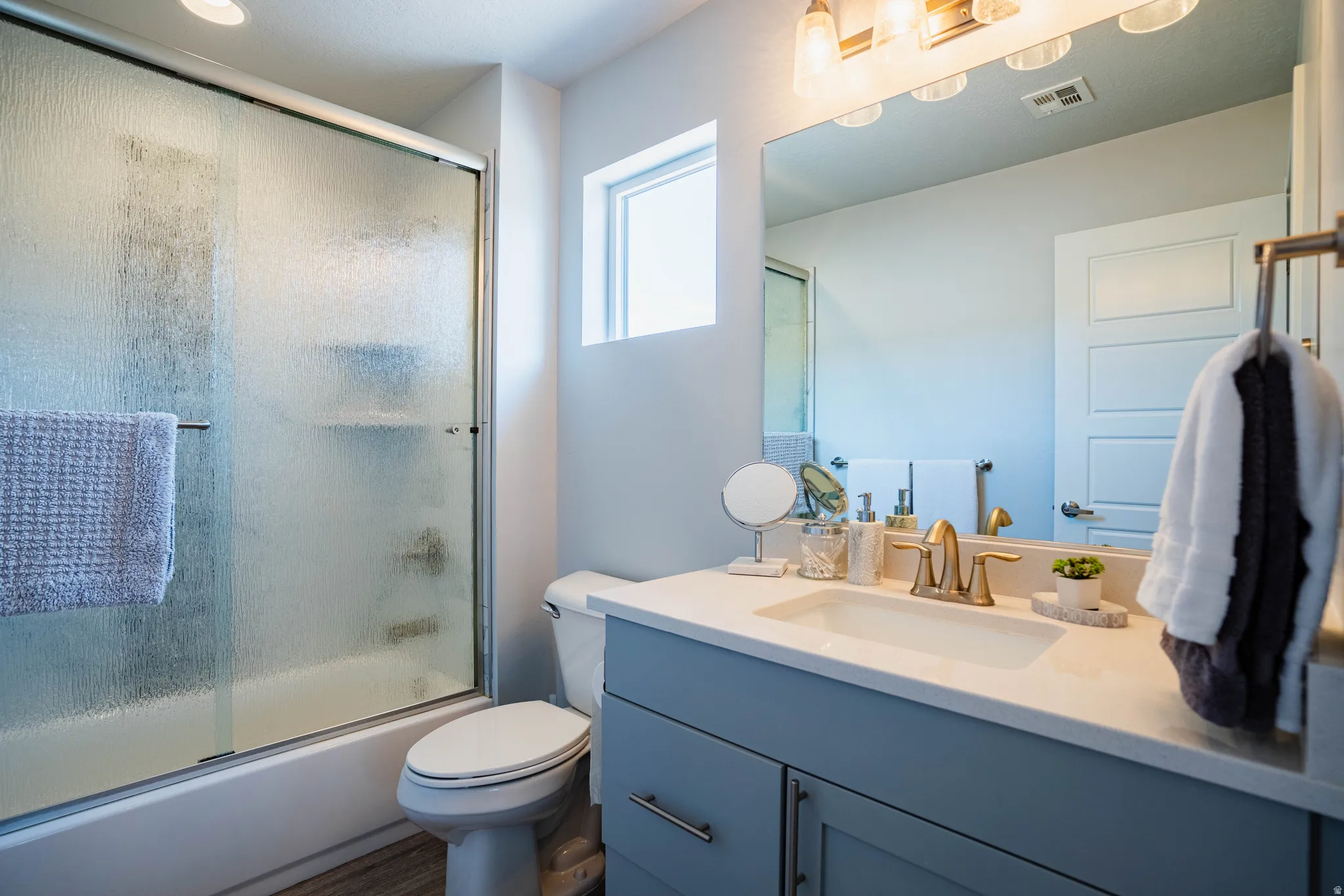 Contemporary bathroom featuring a tub with a sliding glass door, a single vanity with a light countertop, a brass-finish faucet, a flush-mount light fixture, and wood-finish flooring