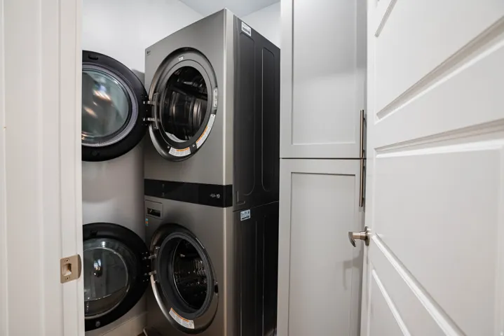 Stacked washer and dryer units with a white paneled door and light-toned cabinetry featuring brushed nickel pulls