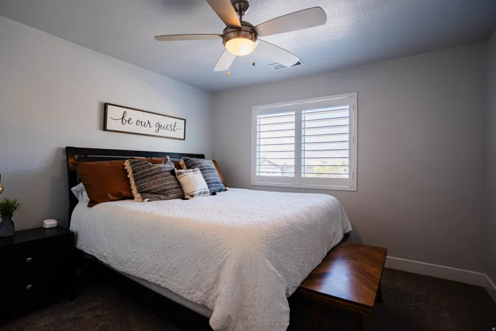 Ceiling fan with integrated lighting, window with white plantation shutters, dark wood-finish bed frame, dark-toned carpet flooring, and light gray wall paint
