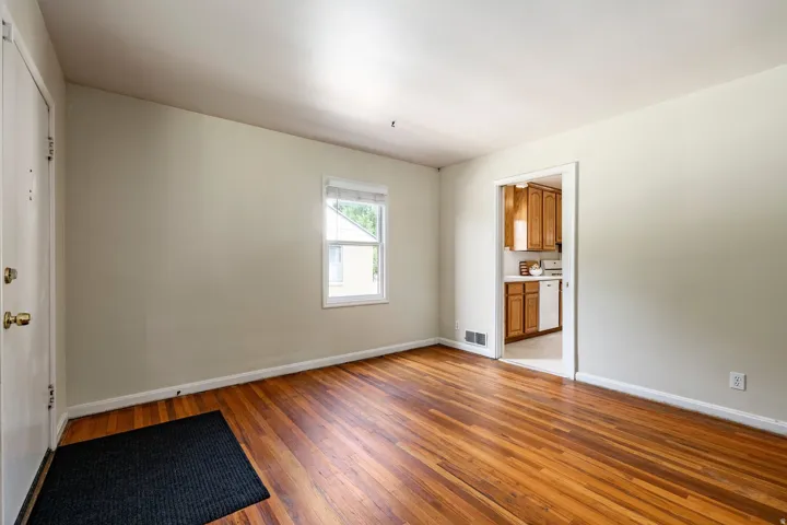 Hardwood flooring throughout, neutral wall tones, single window with white blinds, white trim, and a doorway opening to a kitchen with wood-finish cabinetry