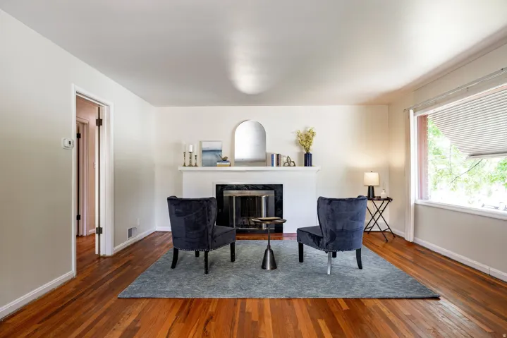 Hardwood flooring throughout, a traditional fireplace with a dark stone surround, and a large picture window with horizontal blinds