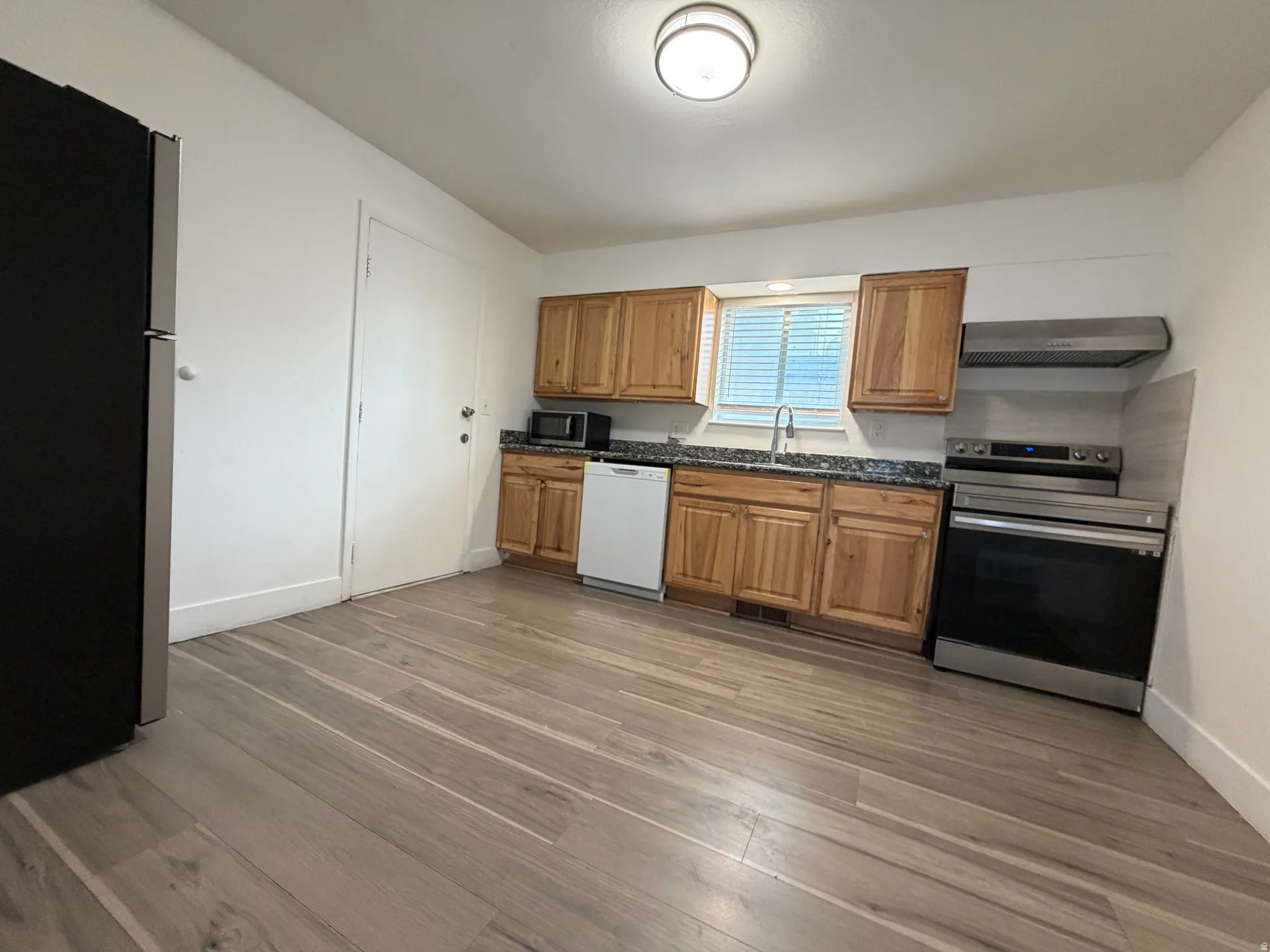 Kitchen featuring wood-finish flooring, natural wood cabinetry, dark countertops, a stainless steel range with matching hood, and a white dishwasher