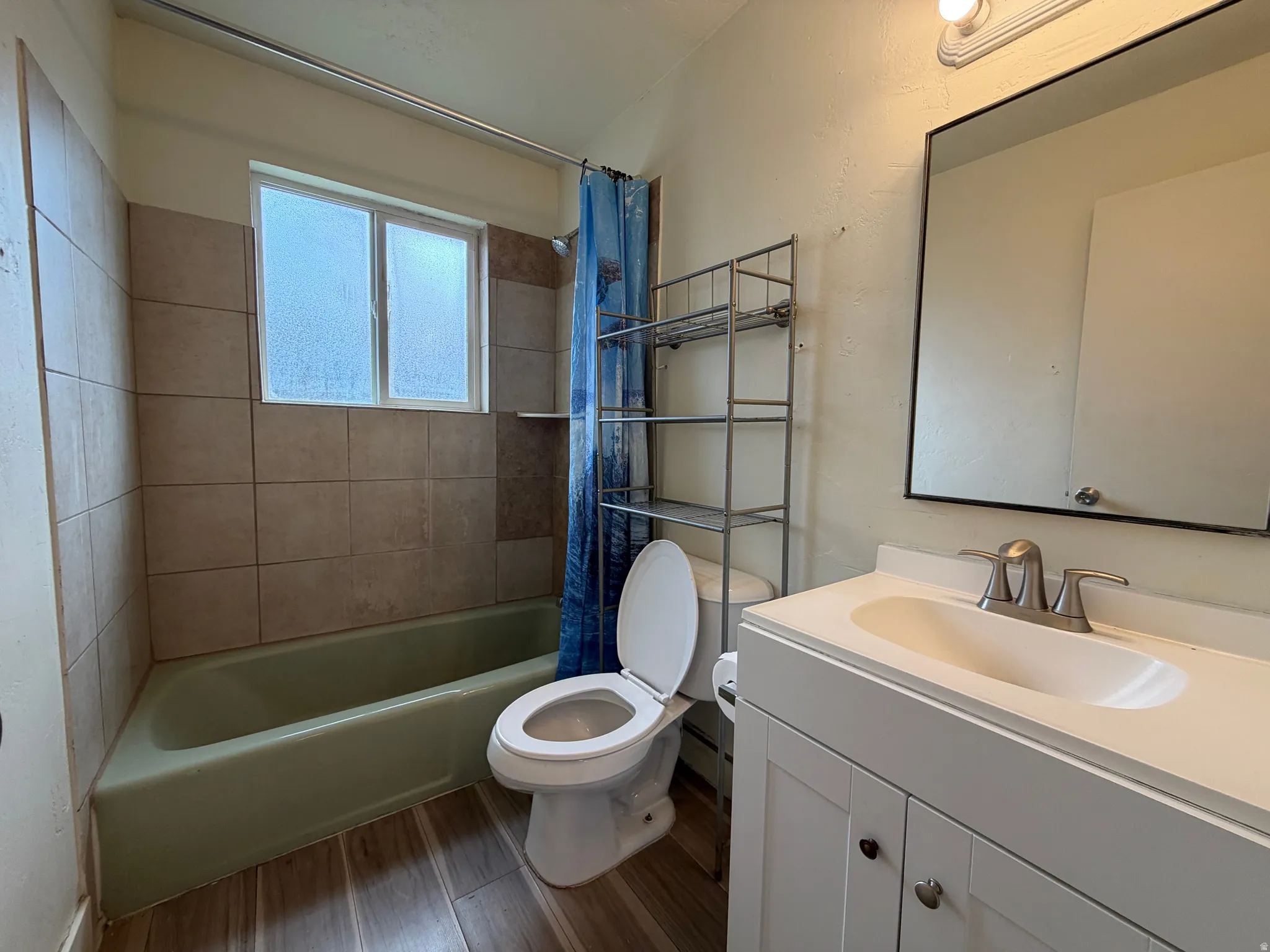 Bathroom featuring a white vanity with integrated sink, brushed nickel faucet, wood-finish flooring, tiled shower surround, and a framed mirror
