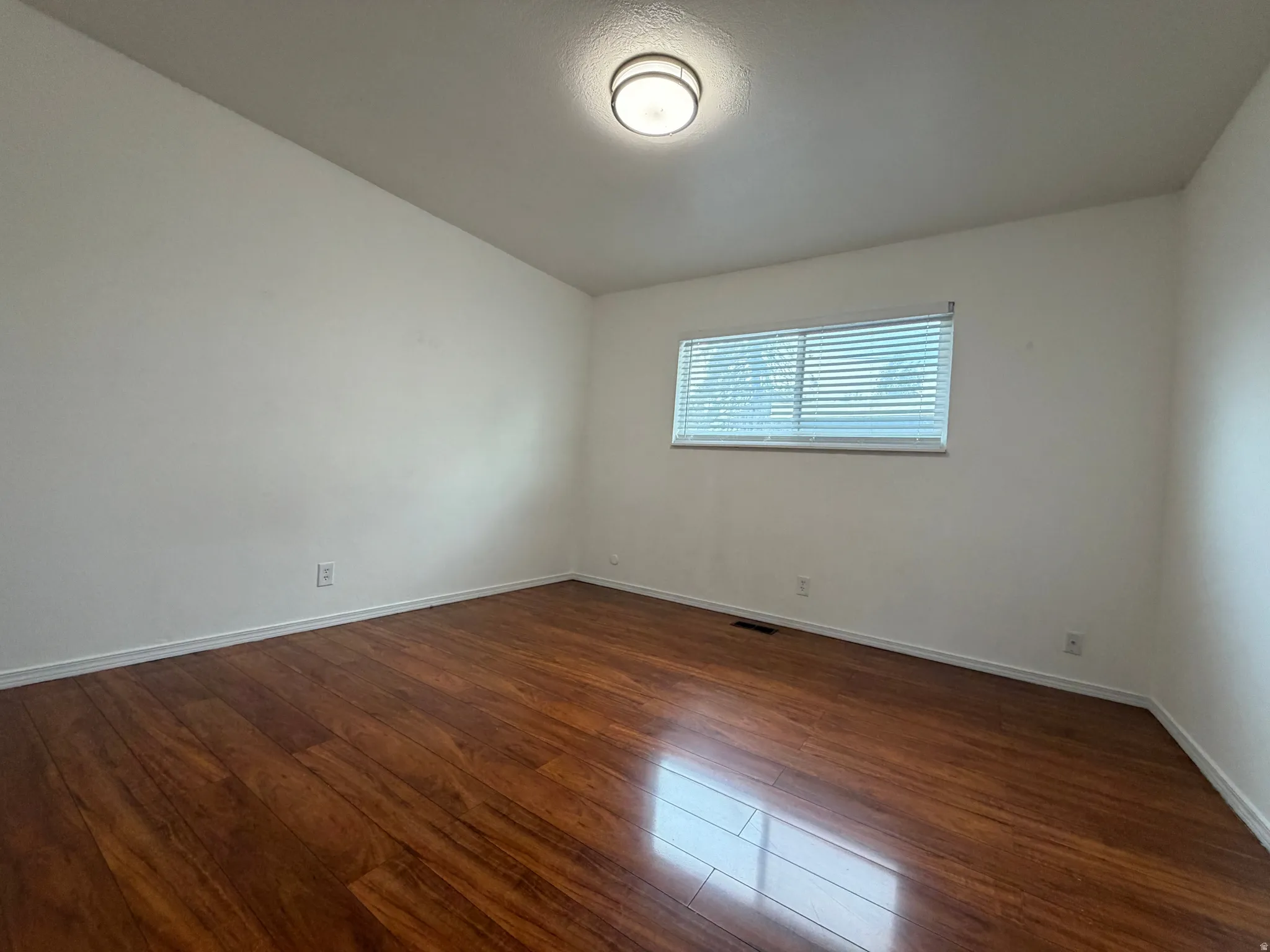 Wood-finish flooring with white walls, a single window with blinds, and a flush-mount ceiling light fixture