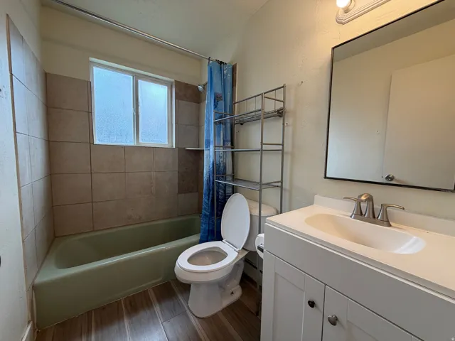 Bathroom featuring a white vanity with integrated sink, brushed nickel faucet, wood-finish flooring, tiled shower surround, and a framed mirror