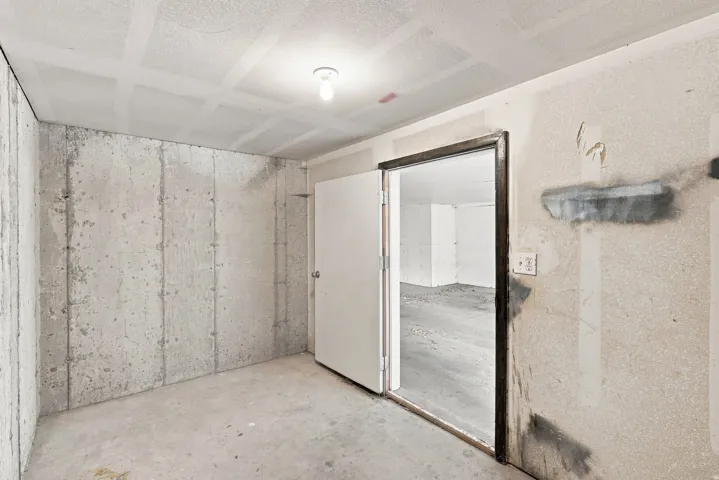 Unfinished concrete walls, unfinished concrete flooring, bare ceiling light fixture, unfinished ceiling, and a white interior door