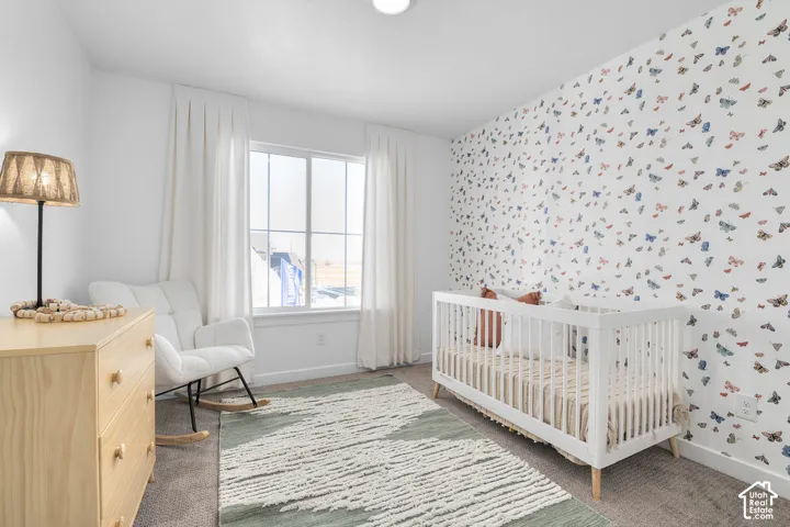 Bright nursery featuring soft patterned wallcovering, large multi-pane window with flowing curtains, white wooden crib, natural wood-finish dresser with integrated lighting, and neutral area rug over carpeted flooring